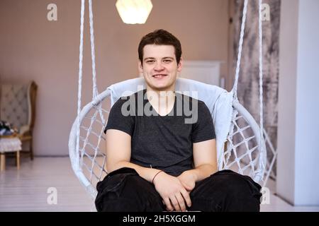 Uomo sorridente di 23 anni in sedia appesa al coperto. Foto Stock