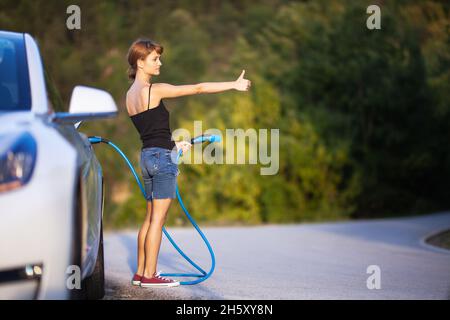 Ragazza in piedi accanto all'auto elettrica tenendo un cavo di ricarica e hitchhiking. Foto Stock