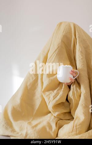 Donna sotto una coperta gialla con la testa. Una tazza con marshmallows e un drink è in mostra all'esterno. Il concetto di autunno e inverno. Foto Stock