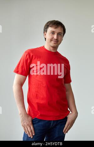 Modello giovane maschile positivo in t shirt rossa casual e jeans guardando la macchina fotografica mentre si è in piedi. Giovane uomo in t shirt rossa su sfondo bianco Foto Stock