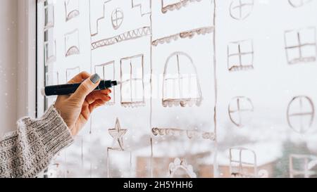 La mano femminile disegna le decorazioni natalizie dipinte su vetro della finestra. Paesaggi di New Years. Edifici della città ghirlande luci. Igge, decorazione e chri Foto Stock