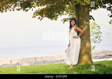 Donna latino con carnagione scura godendosi una passeggiata nel parco lungo il lago, accanto ad un albero. Foto Stock