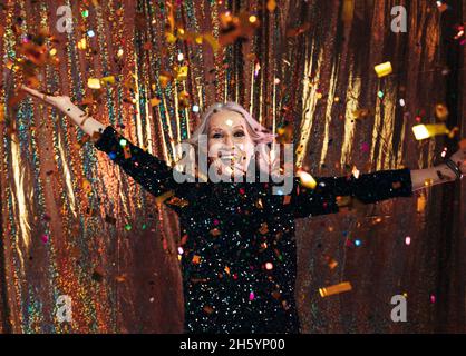 Donna anziana sorridente in abito nero che si diverte sotto i confetti colorati contro uno sfondo dorato Foto Stock