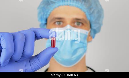 Un bel medico giovane esamina un nuovo farmaco. Un giovane medico bello in una maschera medica tiene una pillola rossa nelle sue mani presentando una nuova droga. Foto Stock