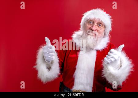 Ritratto Babbo Natale in cappello rosso vestito mostra gesto pollice su come, mostrando pollice su dito. Come su sfondo rosso studio Foto Stock