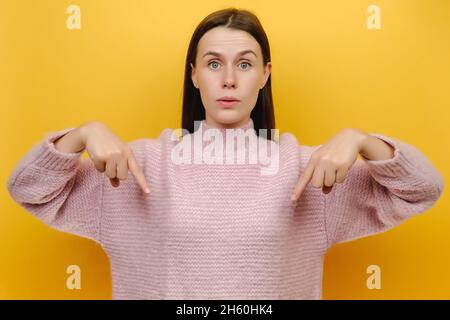 Giovane sorridente felice bella ragazza millennial indossare intimo maglione rosa puntando indice dito giù su area di lavoro copia spazio mockup Foto Stock