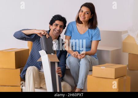 Coppia indiana di mezza età che guarda la macchina fotografica durante il disimballaggio delle scatole nella stanza nuova Foto Stock