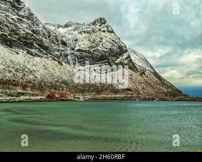 Tipiche case rosse norvegesi sul blu azzurro del mare artico a Ersfjord, con cascate ghiacciate e neve sulle aspre montagne alle spalle. Foto Stock