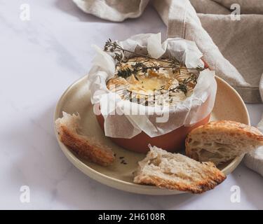 Camembert fonduta con timo in un piccolo piatto di terracotta posto su un piatto da portata con pezzetti di pasta madre. Foto Stock