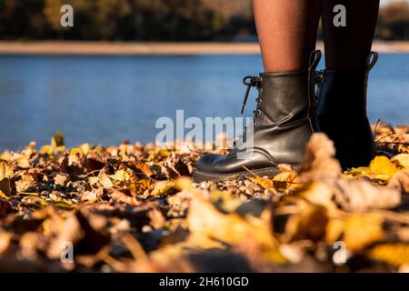 Gambe di donne in pelle nera stivali in piedi sulla spiaggia ricoperta in autunno foglie con lago sullo sfondo Foto Stock