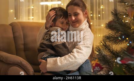 Sorridendo la giovane madre che abbraccia suo figlio seduto in poltrona accanto all'albero di Natale in soggiorno. Famiglie e bambini festeggiano le vacanze invernali.