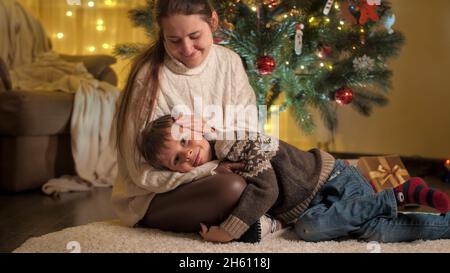 Giovane madre carezzando il figlio sorridente che giace accanto al brillante albero di Natale. Famiglie e bambini festeggiano le vacanze invernali.