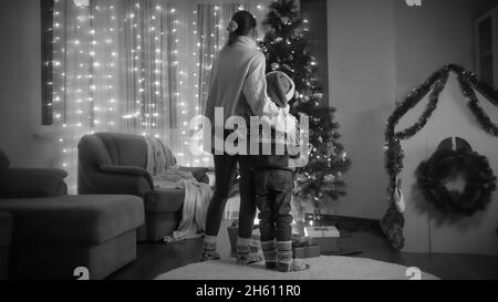 Foto in bianco e nero di mamma con figlio a Santa Hat in piedi accanto al brillante albero di Natale in soggiorno
