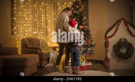 Madre con figlio che festeggia il Natale guardando l'albero di Natale incandescente in soggiorno. Emozioni pure delle famiglie e dei bambini che celebrano l'inverno