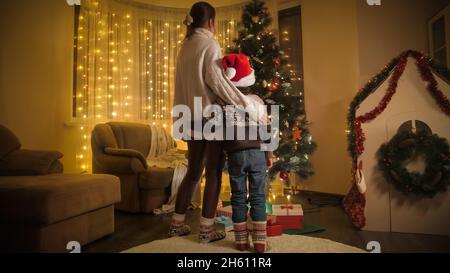 Madre con figlio a Santa Hat in piedi in salotto e guardando su colorato incandescente albero di Natale. Emozioni pure di famiglie e bambini