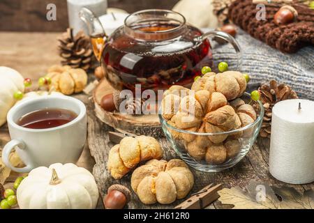 Il concetto di tè d'autunno. Biscotti con purea di zucca, tè nero in teiera di vetro, decorazioni autunnali. Corona, candele, un maglione intimo. Vecchio sfondo in legno, vicino Foto Stock