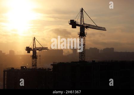 Sagome di gru da costruzione e edifici residenziali incompleti sullo sfondo dell'alba. Costruzione di alloggi, condominio in città Foto Stock