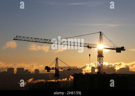 Sagome di gru da costruzione e edifici residenziali incompleti sullo sfondo dell'alba. Costruzione di alloggi, condominio in città Foto Stock