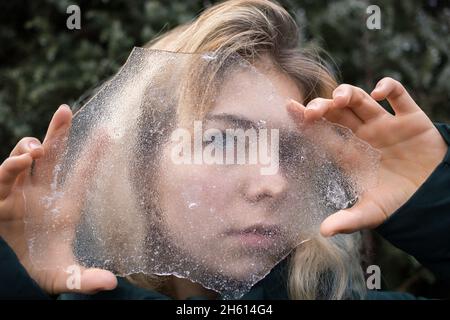 bella giovane donna tiene un grande pezzo di ghiaccio trasparente di fronte al suo viso. Guarda attraverso di esso. Freddo giorno d'inverno Foto Stock