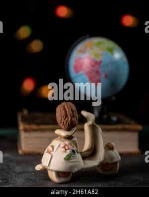 Una noce poggiata su uno scooter in miniatura con un globo su un libro sullo sfondo e bokeh di luci fata colorate Foto Stock