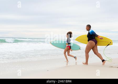 Tutta la lunghezza di spensierate amici femminili multirazziali con tavole da surf che corrono in spiaggia durante il fine settimana Foto Stock