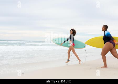 Spensierate amici femminili multirazziali con tavole da surf che corrono in spiaggia durante il fine settimana Foto Stock