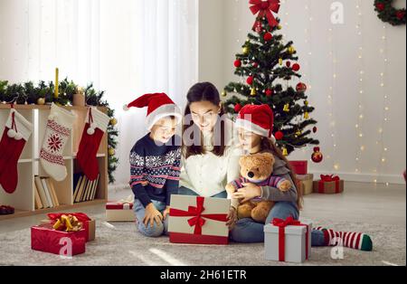 Madre e i suoi bambini piccoli aprono la scatola del regalo di Natale da cui la luce brilla e illumina il viso. Foto Stock