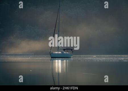Una barca a vela sulle acque calme di una baia riparata che getta riflessi con nebbia mattutina sulla superficie del mare. Foto Stock