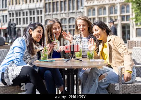 Anversa, Belgio, 21 maggio 2021, quattro turisti o studenti di razza mista seduti fuori nel centro storico della città in una caffetteria terrazza con una videochiamata utilizzando il telefono cellulare. Foto di alta qualità Foto Stock