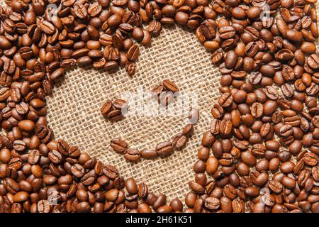 La struttura inclinata a forma di cuore in chicchi di caffè torrefatto con un sorriso su un panno di sackcloth. Foto Stock