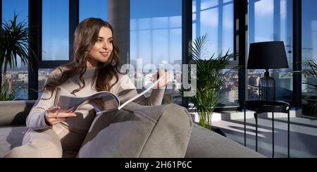 Bella donna con lunghi capelli ondulati rivista lettura mentre si rilassa sul divano. Bella brunetta rilassante in appartamento moderno con finestre panoramiche. Foto Stock