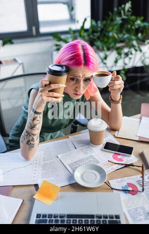 vista ad angolo di una donna d'affari stressata con capelli rosa che tengono il caffè e guardano il computer portatile Foto Stock