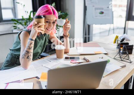 donna d'affari preoccupata con i capelli rosa che tengono il caffè e guardando il computer portatile Foto Stock