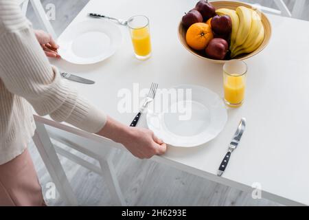 Vista ritagliata della donna mettendo piatti sul tavolo vicino succo d'arancia e frutta in cucina Foto Stock