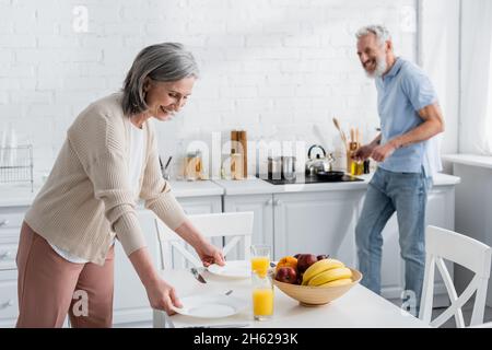 Donna allegra matura mettendo piatti vicino succo d'arancia ad un marito sfocato in cucina Foto Stock