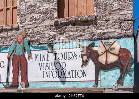 Tonopah, Nevada, Stati Uniti - 16 maggio 2011: Primo piano di cartello Dowtown dipinto di colore per i tour delle miniere con uomo anziano e asino contro il muro di pietra grigia. Foto Stock