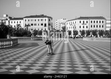 un ragazzo e una ragazza pattinano insieme su uno skateboard sulla terrazza mascagni di livorno, italia, un ampio belvedere sinuoso verso il mare con una superficie di pavimentazione di 8700 mq a scacchiera e 4,100 balaustre Foto Stock