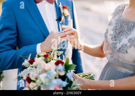 Sposa in un abito in pizzo mette un anello di aggancio sul dito di uno sposo in un abito blu. Primo piano Foto Stock