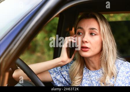 Giovane donna che parla su un telefono cellulare mentre è seduto in auto sul sedile del conducente. Attraente studentessa bionda femminile. Foto Stock