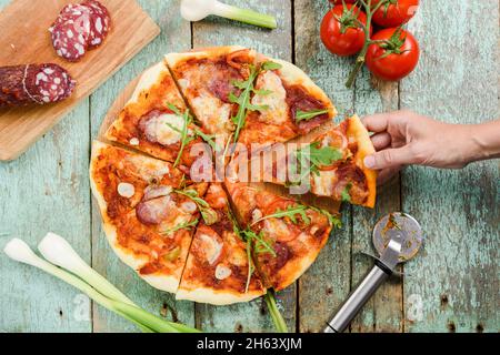 Pizza al forno fatta a mano con salame affumicato, pomodori, olive verdi e formaggio serviti con coltello da pizza e ingredienti: Porri, pomodori ciliegini e salumi. Foto Stock