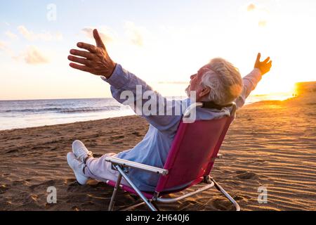 un vecchio e maturo che si gode le vacanze estive da solo sulla spiaggia seduta in una piccola sedia che guarda al mare. uomo che si sente libero con le braccia aperte. concetto di libertà. Foto Stock