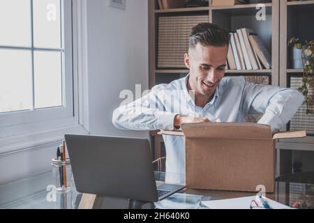 felice uomo sorridente che lavora e apre una piccola scatola appena consegnata a casa godendo la sua sorpresa da solo divertirsi. giovane maschio caucasico che sboxing un po 'di regalo o regalo in ufficio. Foto Stock