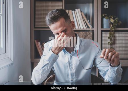 malsano stressato uomo d'affari che tollera gli occhiali, le palpebre di sfregamento, che soffrono di sindrome degli occhi asciutti a causa di lavoro lungo del calcolatore, il ponte della testa massaggiante che allevia il dolore in ufficio a casa. Foto Stock