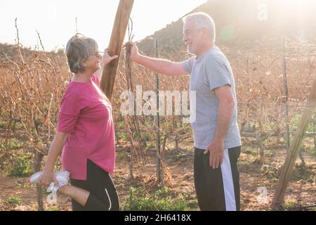 felice coppia attiva di persone anziane fare esercizi di stretching all'aperto insieme sorridente - giovane anziano stile di vita uomo e donna in pensione in attività sportiva con vigneto in background Foto Stock