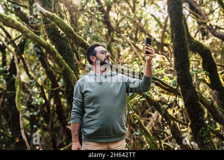 uomo maturo in occhiali che prende selfie o video chattando con il telefono cellulare in foresta. turista maschio che cerca il segnale con uno smartphone in natura. uomo che cerca la rete utilizzando il telefono in foresta. Foto Stock