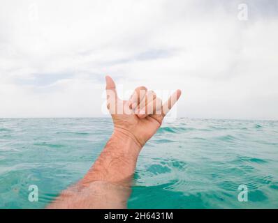 primo piano della mano dell'uomo che mostra il segno shaka sopra l'acqua di mare contro il cielo. mano di un uomo che esce dalla superficie dell'acqua di mare e che gestura il segno shaka contro il cielo nuvoloso Foto Stock