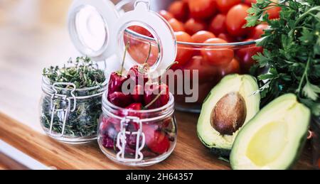 primo piano di peperoncini rossi secchi ed erbe in vaso con verdure biologiche fresche in ciotola. spezie e verdure come carota, lattuga, pomodori con avocado sul banco da cucina per preparare il cibo. Foto Stock