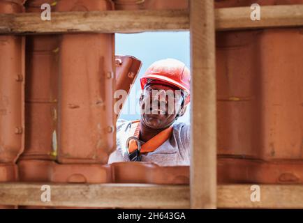 Operatore latino che installa tegole gialle in ceramica montate su tavole di legno che coprono il tetto di edifici residenziali in costruzione. Foto Stock