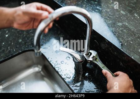 Primo piano delle mani di vecchio riparatore che fissa il rubinetto rotto della cucina usando la chiave regolabile. Concetto di servizio di riparazione. Foto Stock