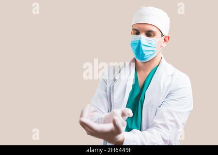 Giovane medico in tuta bianca e maschera protettiva per la respirazione mette i guanti sulle mani su sfondo giallo isolato. Foto Stock